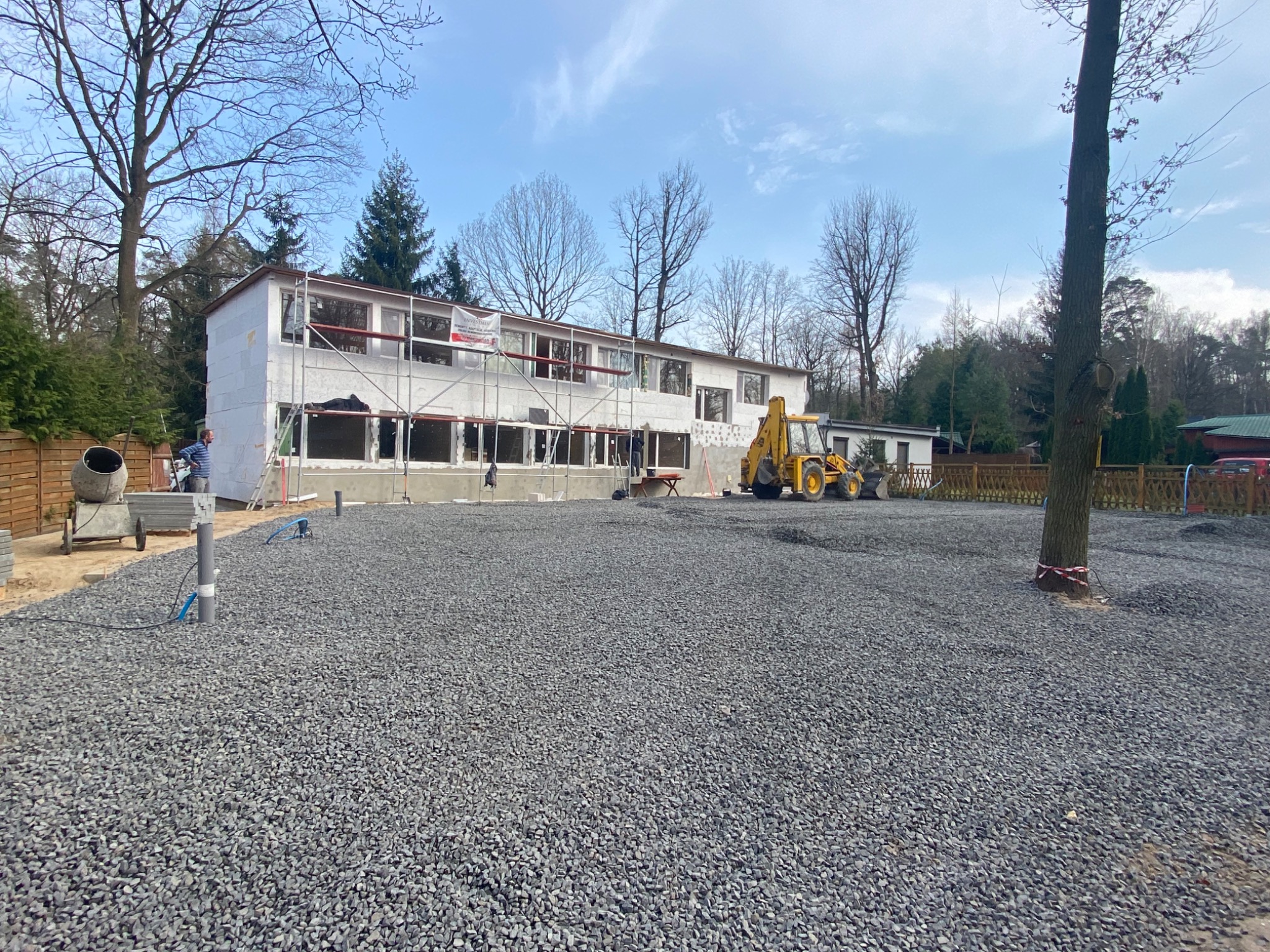 Dwukondygnacyjny budynek w trakcie ocieplania elewacji, widoczne rusztowania, mieszalnik betonu i koparka na placu budowy z wysypanym szarym gruzem.