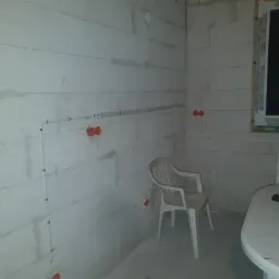 Wnętrze w trakcie prac instalacyjnych: widoczne przewody elektryczne prowadzone po ścianie z betonu komórkowego, puszki instalacyjne, plastikowe krzesło i stół z roboczymi rękawicami.