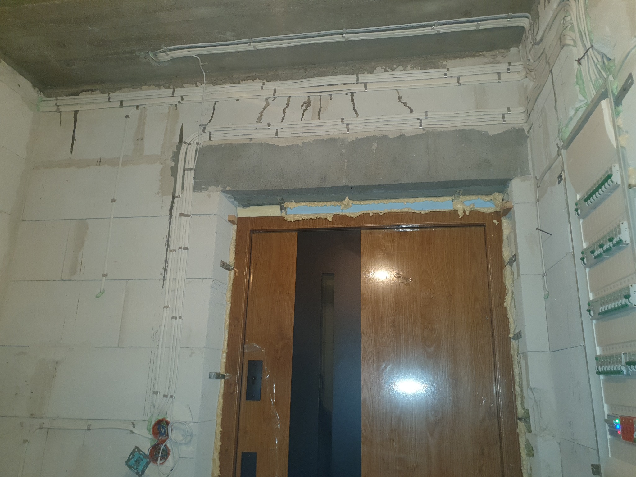 Instalacja elektryczna w trakcie realizacji: kable prowadzone wzdłuż sufitu i ścian, puszki elektryczne wystają ze ściany, widoczna skrzynka rozdzielcza na ścianie obok futryny drzwiowej.