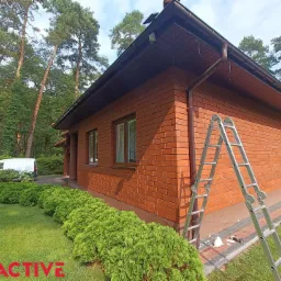 Dom z cegły klinkierowej z brązowymi rynnami, drabina oparta o ścianę, widoczny fragment instalacji. Na trawniku leżą narzędzia. W tle las i biały samochód dostawczy.