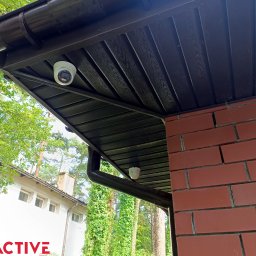 EXEACTIVE - Dwie kamery monitoringu zamontowane pod brązowym dachem domu z ceglaną ścianą. Widok z dołu na tle zieleni.