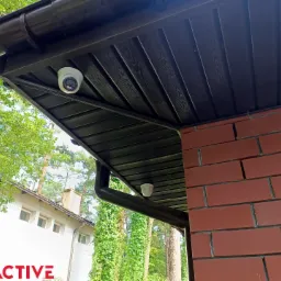 Dwie kamery monitoringu zamontowane pod brązowym dachem domu z ceglaną ścianą. Widok z dołu na tle zieleni.