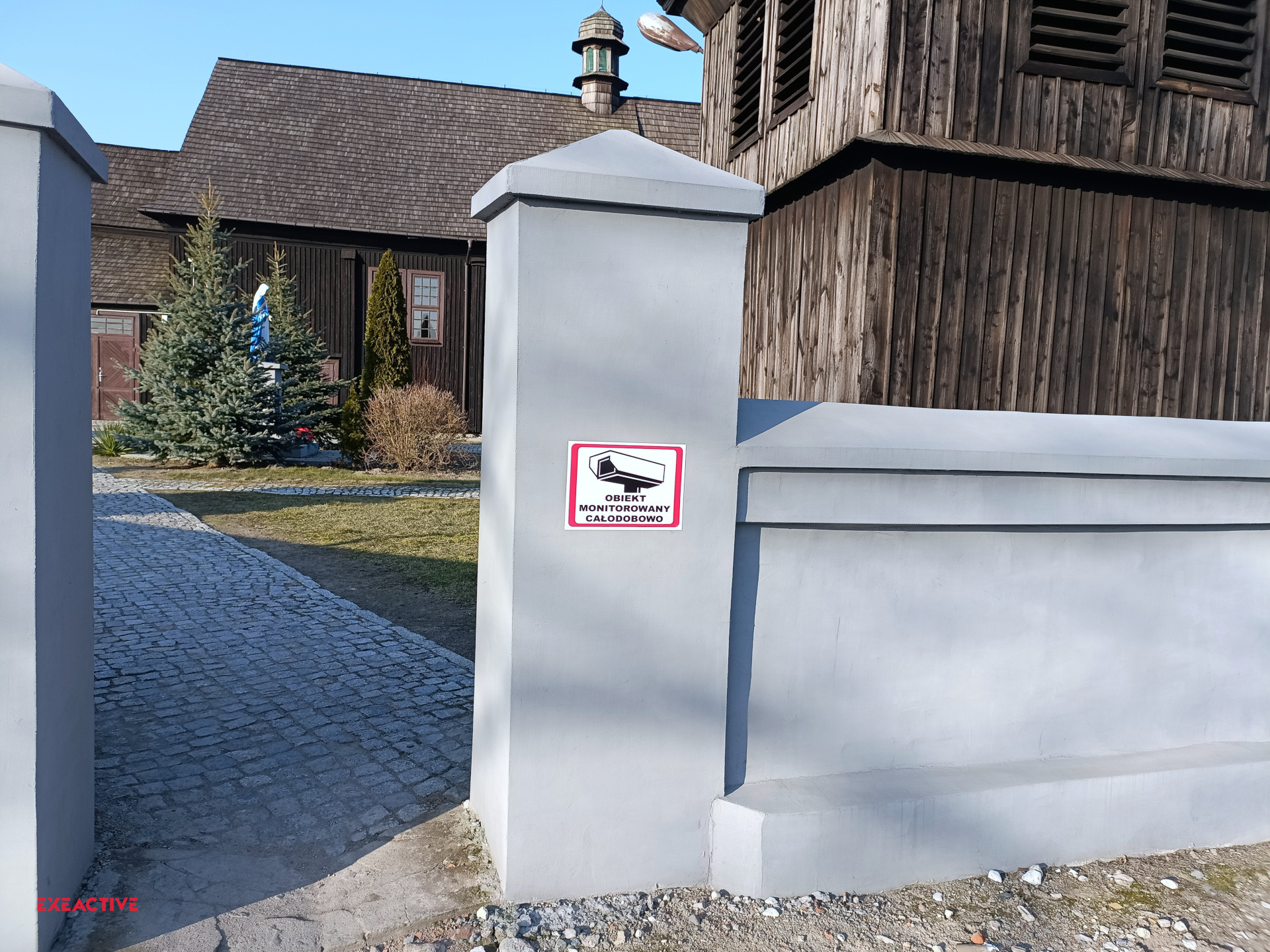 Tabliczka informacyjna 'Obiekt monitorowany całodobowo' umieszczona na szarym słupku ogrodzeniowym, w tle drewniany kościół i chodnik z kostki brukowej.