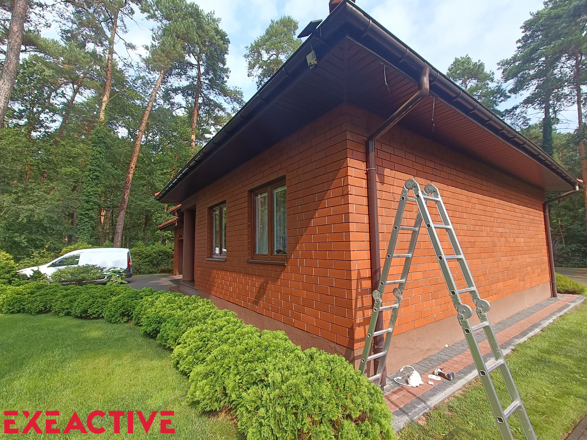 Dom z cegły klinkierowej z brązowymi rynnami, drabina oparta o ścianę, widoczny fragment instalacji. Na trawniku leżą narzędzia. W tle las i biały samochód dostawczy.
