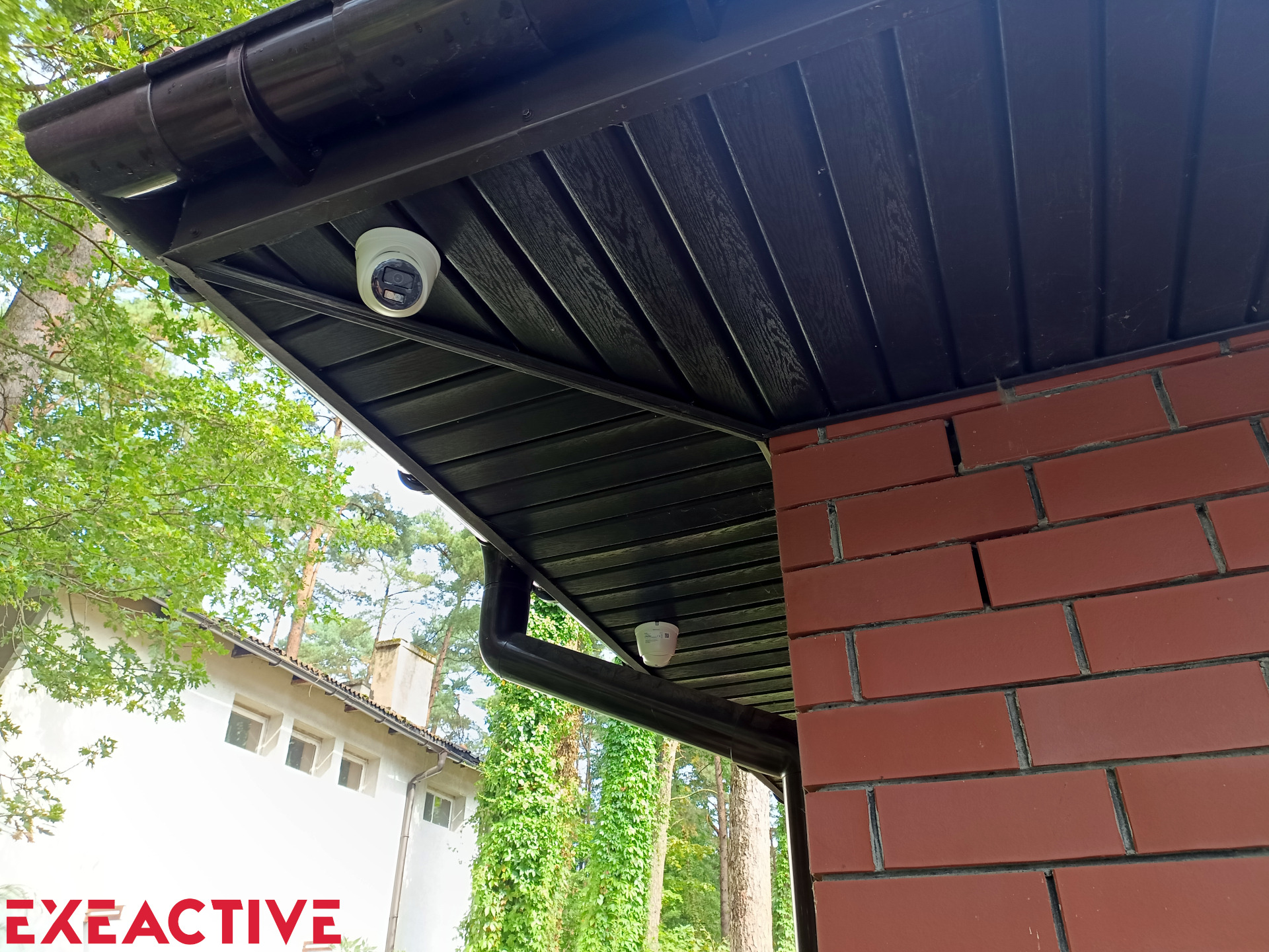 Dwie kamery monitoringu zamontowane pod brązowym dachem domu z ceglaną ścianą. Widok z dołu na tle zieleni.