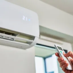 Biały klimatyzator ścienny z wyświetlaczem ustawionym na 21 stopni, sterowany pilotem trzymanym w dłoni, widoczne żaluzje wylotowe.