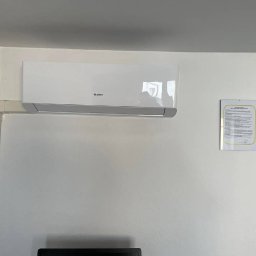 Lispo Mateusz Kleniewski - Biały klimatyzator marki Gree zamontowany na ścianie, widoczna instalacja elektryczna w białej listwie i kamera monitoringu w rogu pomieszczenia, obok wisi dokument w ramce.