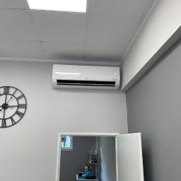 Lispo Mateusz Kleniewski - Biały klimatyzator ścienny marki LG zamontowany w biurze, obok ozdobny zegar z rzymskimi cyframi i widoczne otwarte drzwi do innego pomieszczenia.