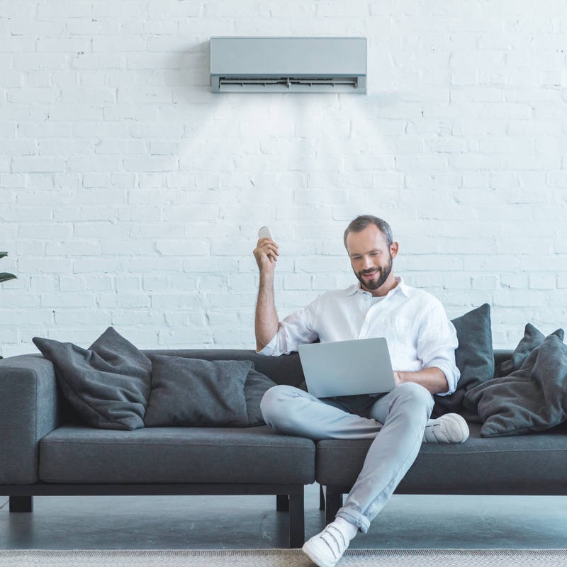 Mężczyzna z brodą siedzi na szarej sofie z laptopem i pilotem klimatyzacji w ręku, pod ścianą z białą cegłą, na której zamontowany jest klimatyzator.