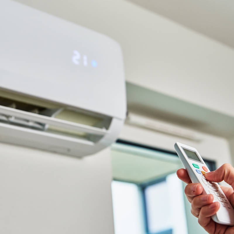 Biały klimatyzator ścienny z wyświetlaczem ustawionym na 21 stopni, sterowany pilotem trzymanym w dłoni, widoczne żaluzje wylotowe.