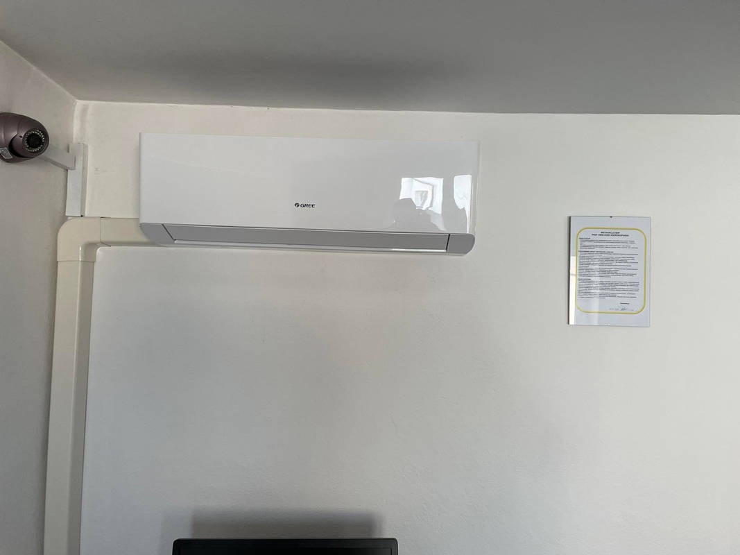 Biały klimatyzator marki Gree zamontowany na ścianie, widoczna instalacja elektryczna w białej listwie i kamera monitoringu w rogu pomieszczenia, obok wisi dokument w ramce.