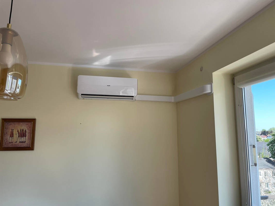Biała klimatyzacja AUX zamontowana na ścianie w jasnym pokoju, z widocznymi białymi listwami maskującymi rury, obrazek na ścianie i lampa wisząca w kadrze.