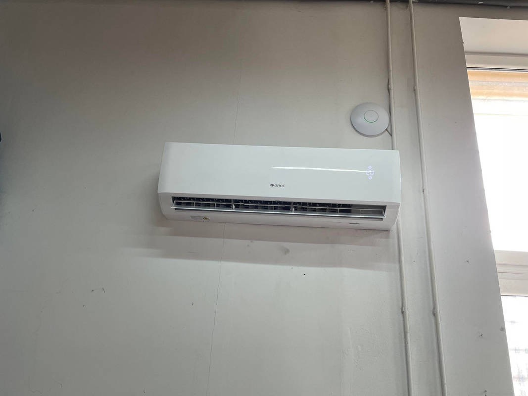 Biały klimatyzator marki Gree zamontowany na jasnej, pomalowanej ścianie w pomieszczeniu, obok widoczny punkt dostępowy Wi-Fi.