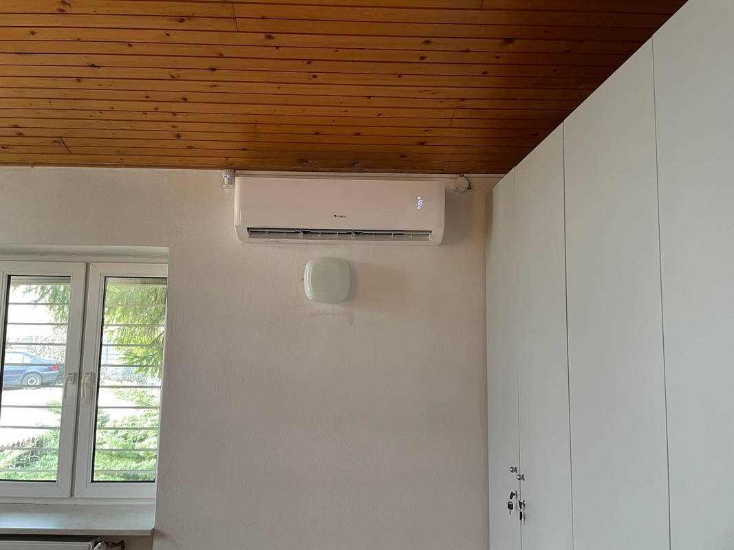 Biały klimatyzator zamontowany na ścianie w pokoju z drewnianym sufitem, obok okna i białej szafy.