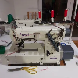 Dwie przemysłowe maszyny do szycia overlock marki Texi z nawleczonymi nićmi w odcieniach czerwieni i zieleni, obok leżą złote nożyczki krawieckie.