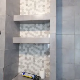 Nowoczesna łazienka z wnęką wyłożoną mozaiką w odcieniach szarości, dwie półki z płytek, tuba silikonu z pistoletem i rolka papieru toaletowego na dolnej półce.