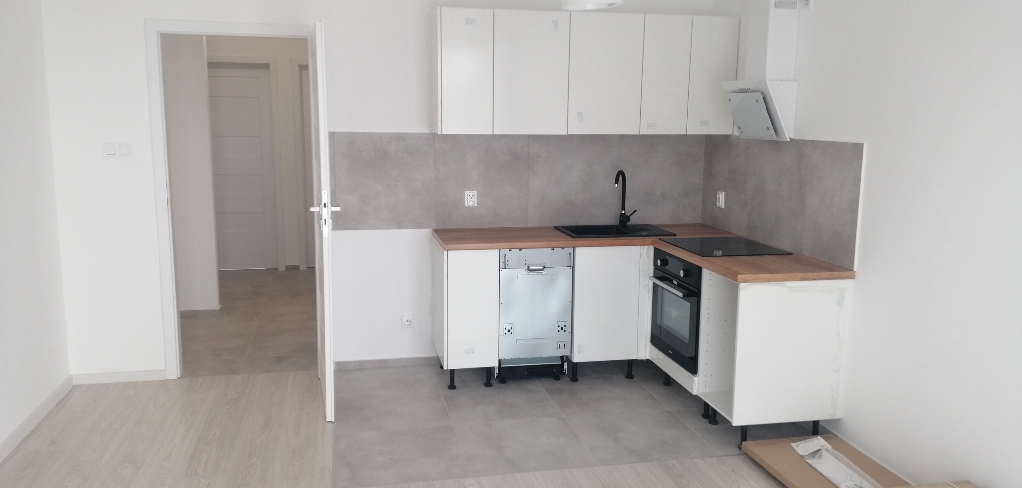 Nowoczesna, minimalistyczna kuchnia z białymi szafkami, drewnianym blatem, czarnym zlewem i płytą indukcyjną, w trakcie instalacji, widoczne niedociągnięcia wykończeniowe i otwarte drzwi do innego...
