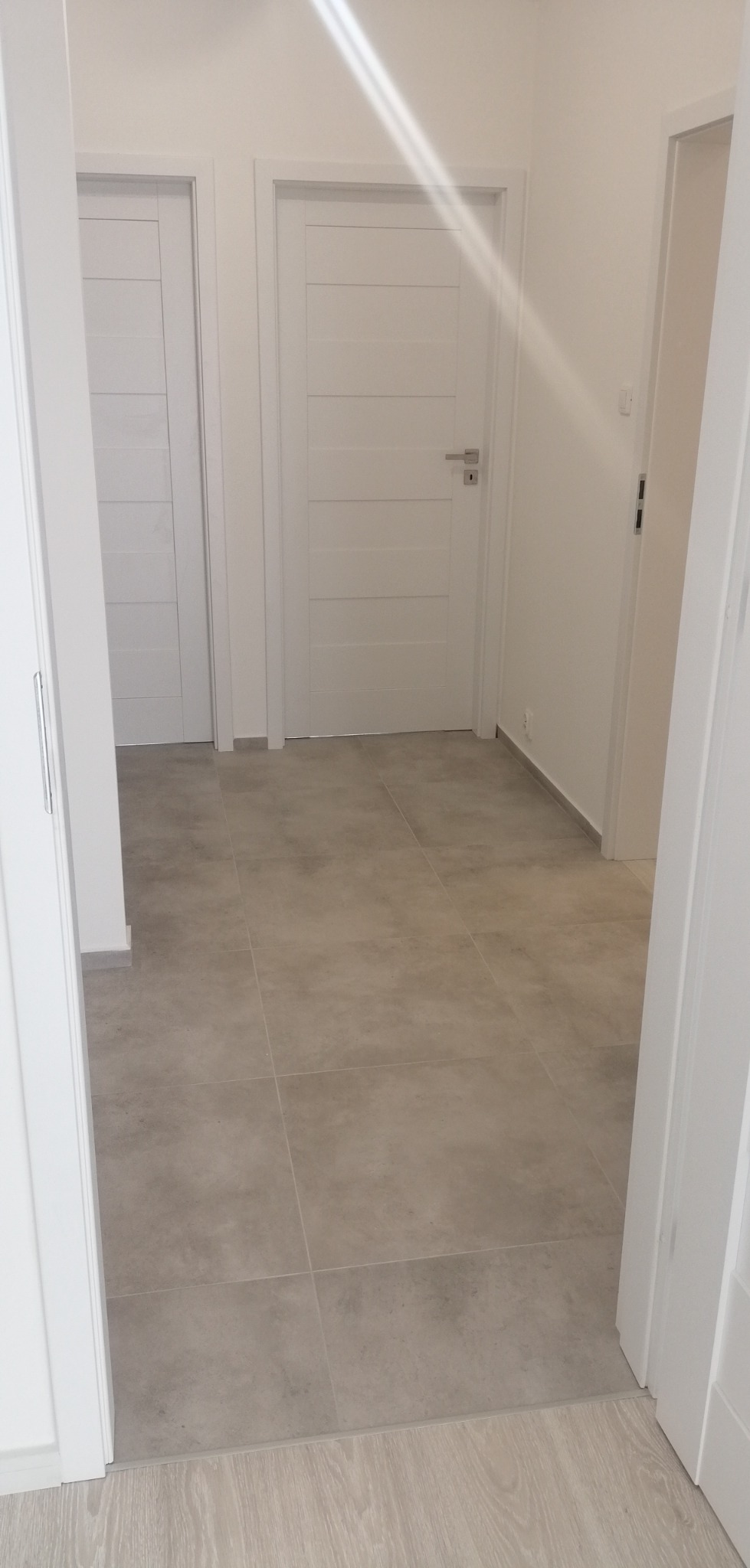 Jasne płytki podłogowe w korytarzu z białymi drzwiami wewnętrznymi z poziomymi frezami i srebrnymi klamkami.