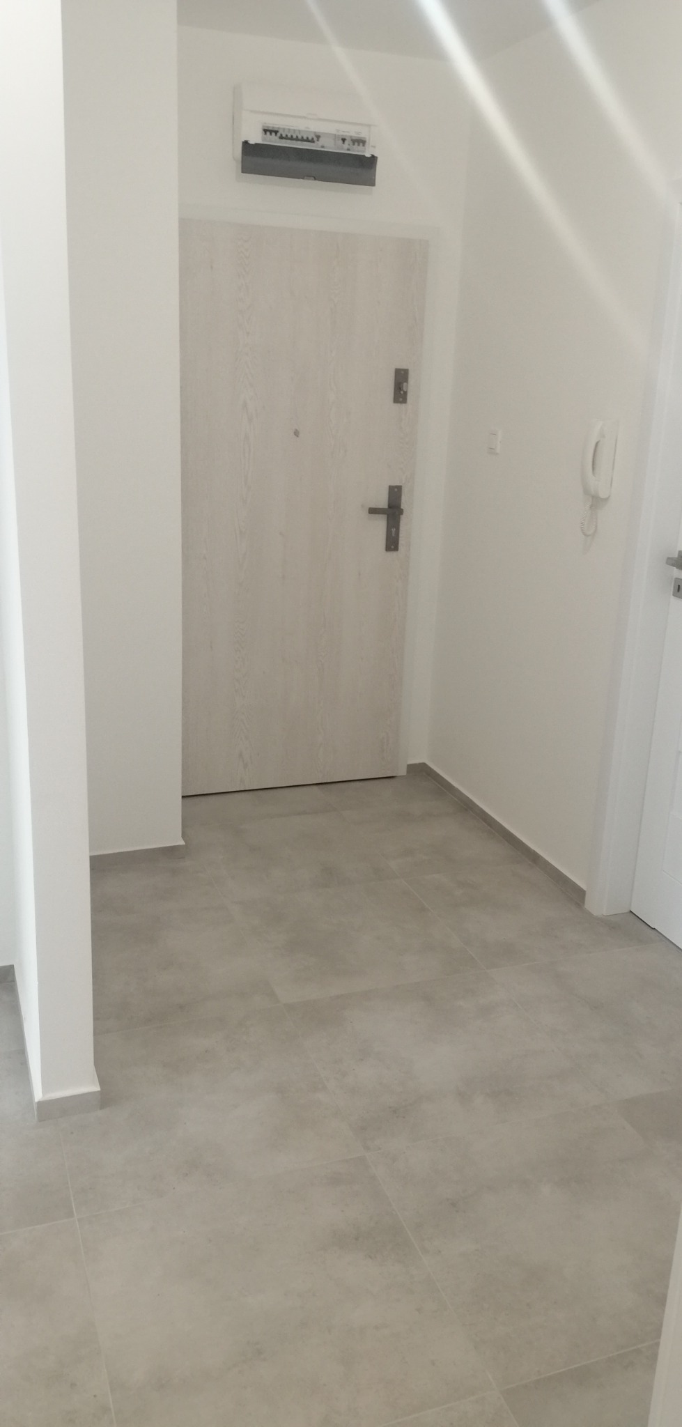 Wnętrze po remoncie: szare płytki podłogowe, białe ściany, drzwi wejściowe z jasnego drewna i skrzynka elektryczna nad nimi.