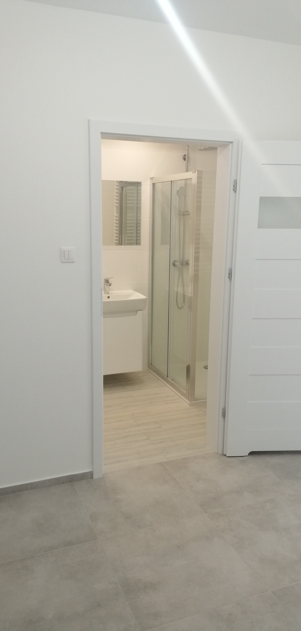 Widok na świeżo wyremontowaną łazienkę z kabiną prysznicową i umywalką, widoczną przez otwarte białe drzwi. Podłoga wyłożona płytkami w dwóch odcieniach szarości.