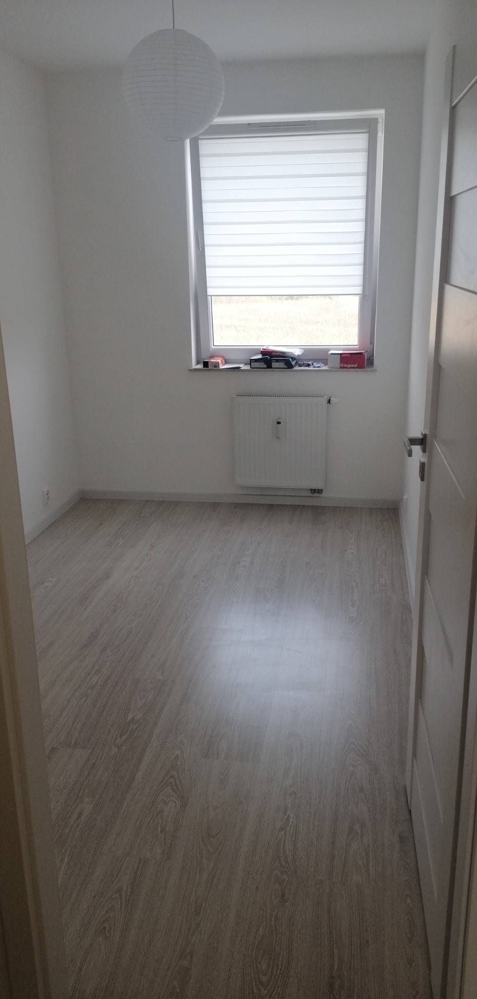 Pusty, jasny pokój z panelami podłogowymi w odcieniach szarości, białymi ścianami, grzejnikiem pod oknem z roletą i wiszącą lampą kulistą.