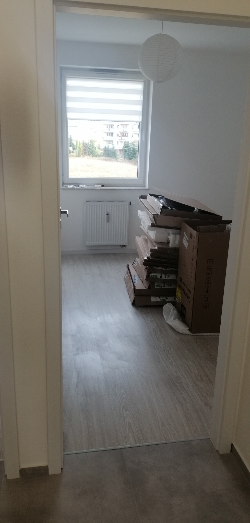 Pusty pokój z nową podłogą z paneli, sterta kartonów po sprzęcie AGD przy ścianie, widok z korytarza przez otwarte drzwi.