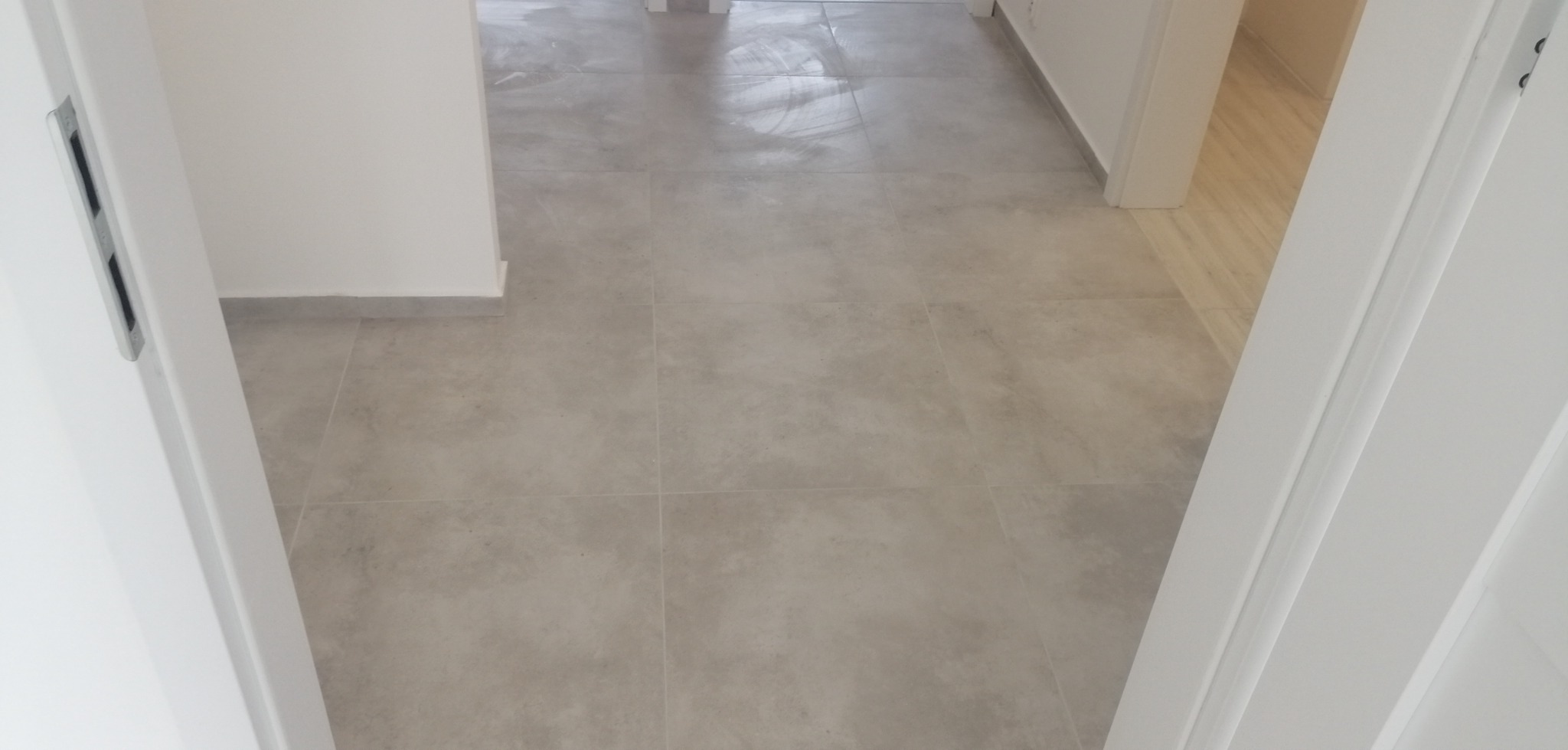 Szare płytki podłogowe ułożone w nowym mieszkaniu, widok z korytarza przez białe futryny drzwiowe.