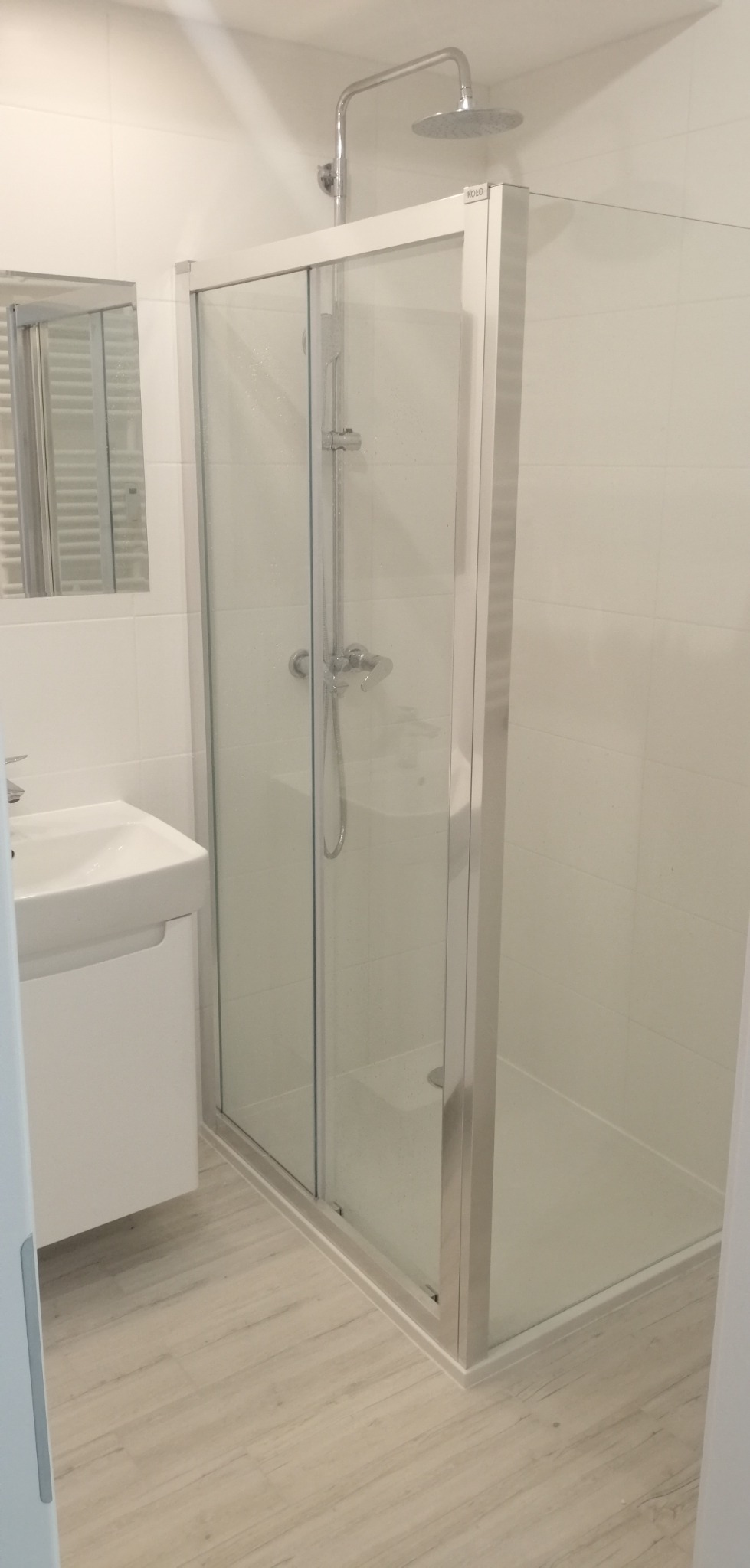 Nowoczesna, przeszklona kabina prysznicowa z przesuwnymi drzwiami, zamontowana w jasnej łazience z białą umywalką i lustrem.