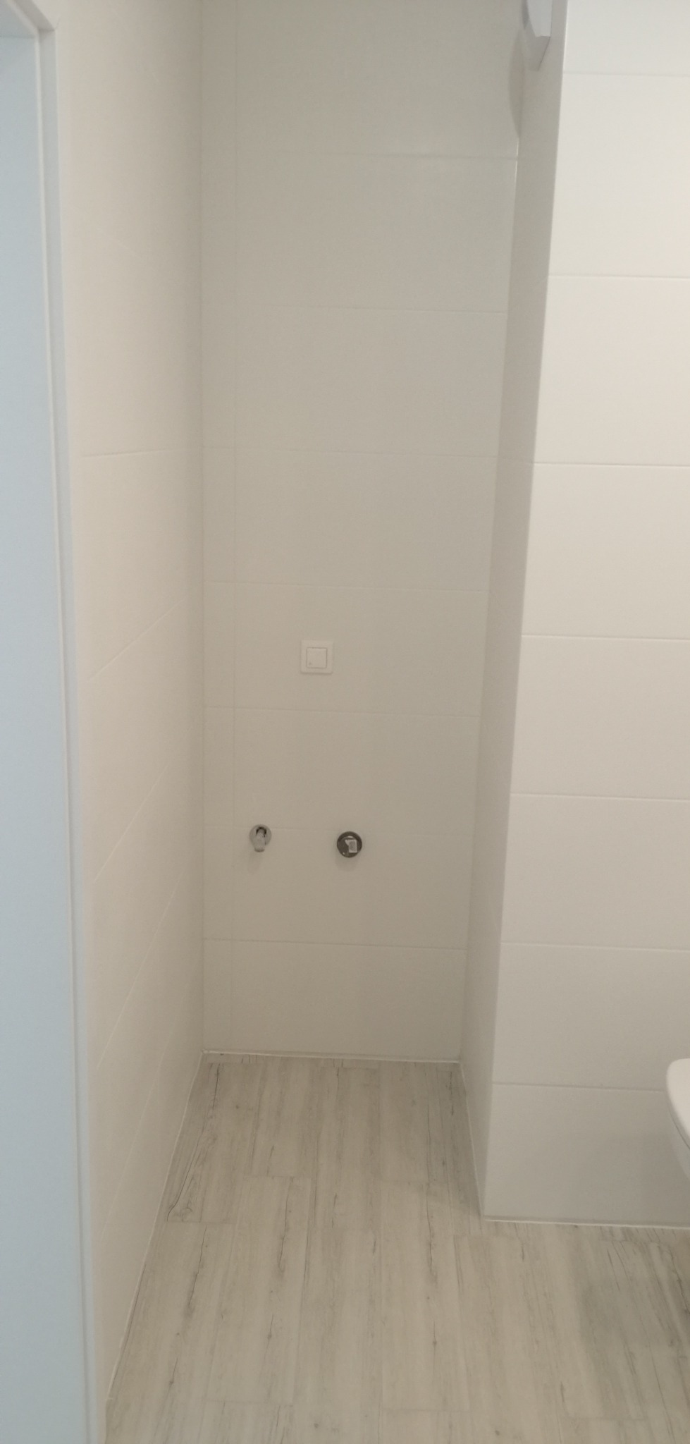 Świeżo wykafelkowana, jasna łazienka z panelami podłogowymi imitującymi drewno, widoczne przyłącza wodne i włącznik światła, brak armatury.