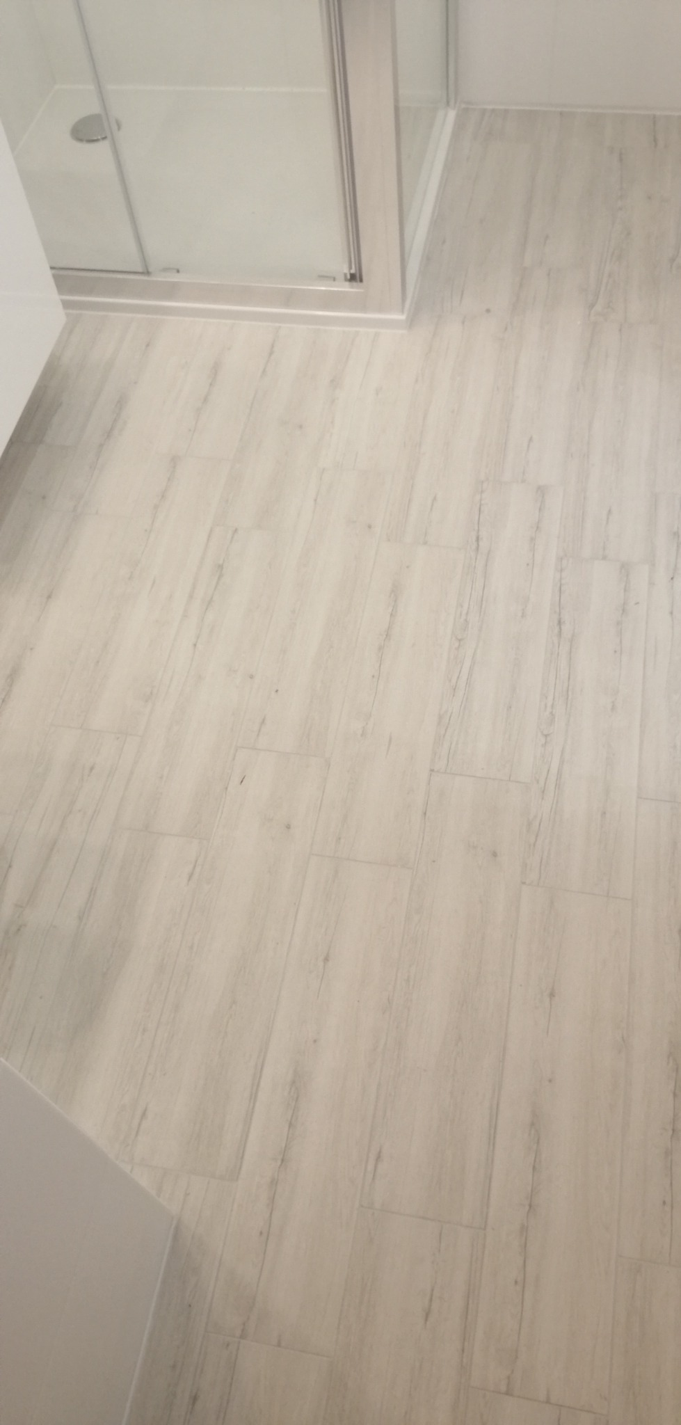 Jasne panele podłogowe w łazience z kabiną prysznicową, imitujące drewno, ułożone ukośnie.