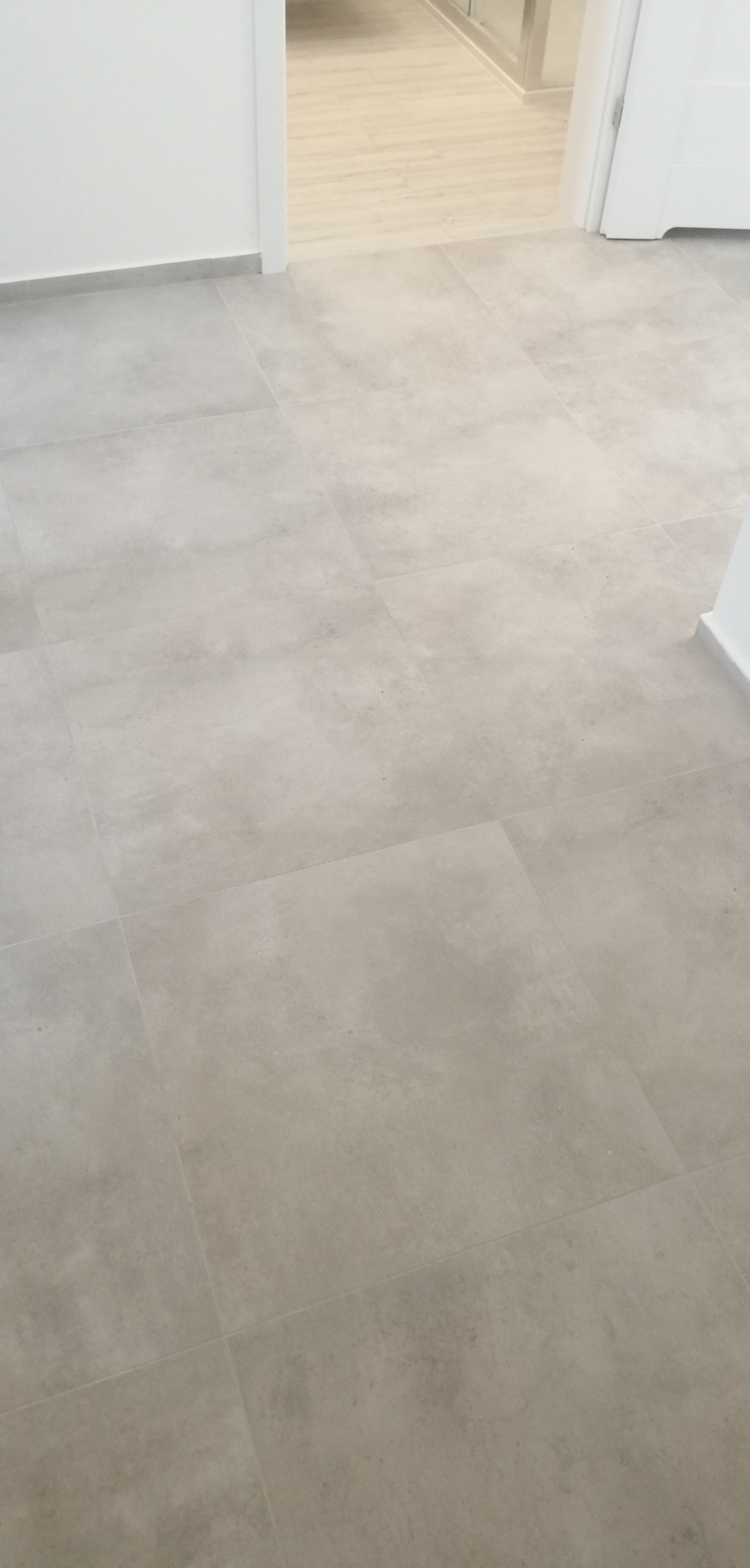 Szare płytki podłogowe o matowej powierzchni, ułożone w kwadraty, widoczne przejście do pomieszczenia z jasną drewnianą podłogą, biała listwa przypodłogowa.
