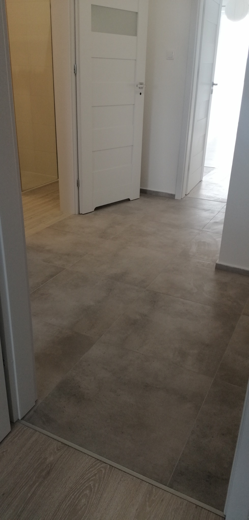 Wykończony korytarz z szarą, matową podłogą z płytek, białe drzwi wewnętrzne z przeszkleniami, widoczne przejście podłogi z płytek na panele.