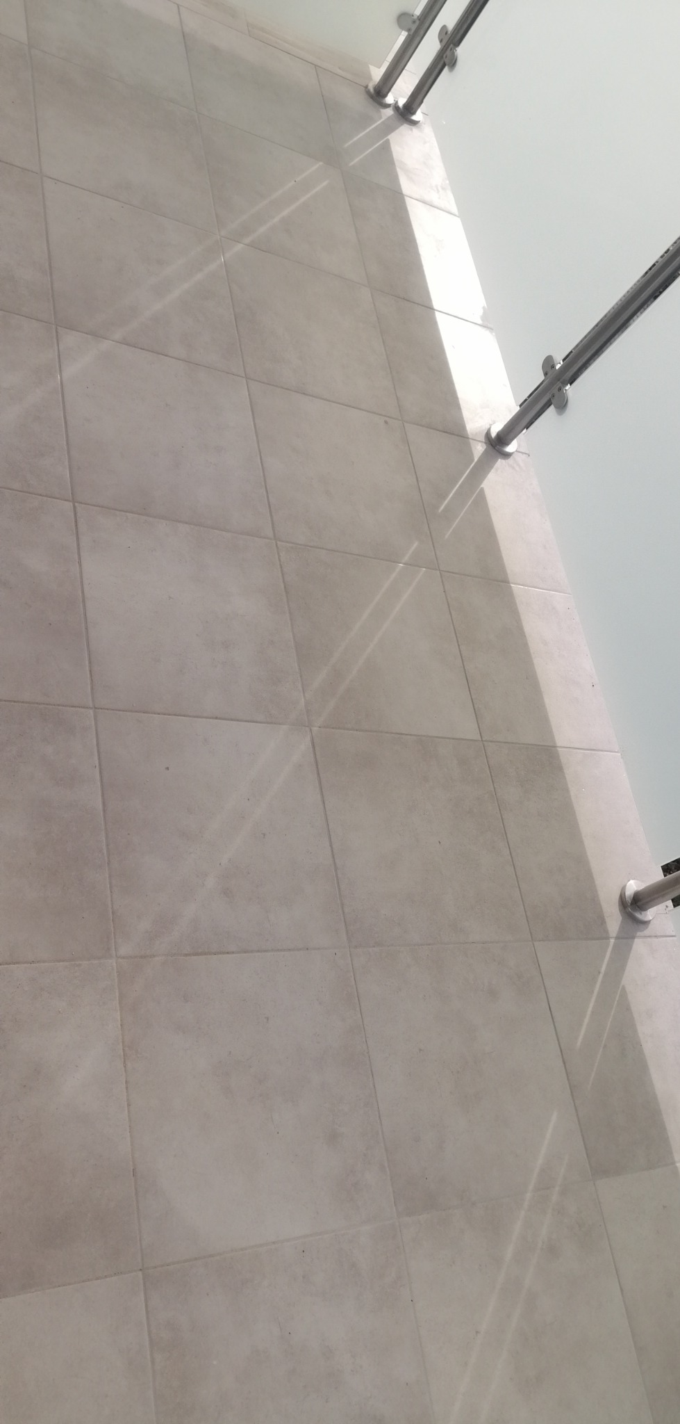 Jasne, kwadratowe płytki podłogowe w neutralnym odcieniu, z widocznymi cieniami rzucanymi przez metalową balustradę balkonu, ukazujące ukończoną powierzchnię.