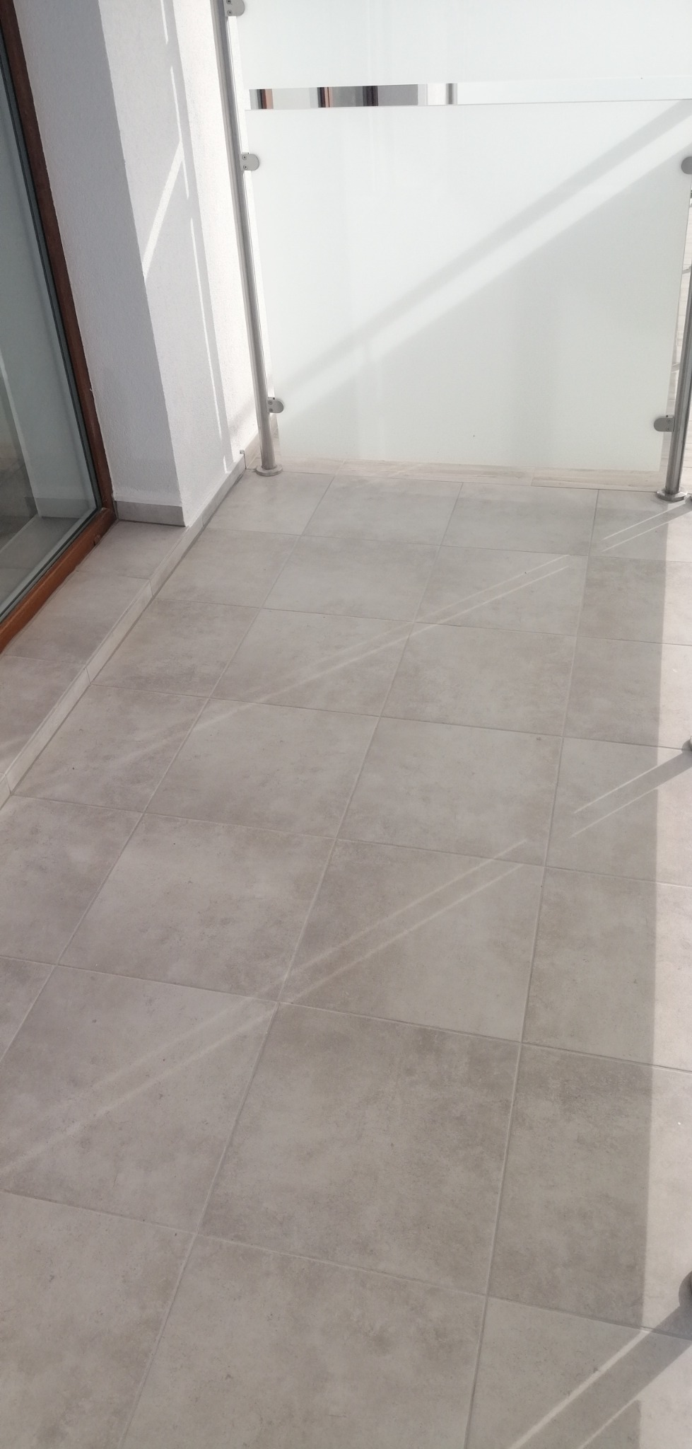 Wykończony balkon z jasnymi płytkami podłogowymi i szklaną balustradą z metalowymi elementami mocującymi, widoczne refleksy słoneczne.