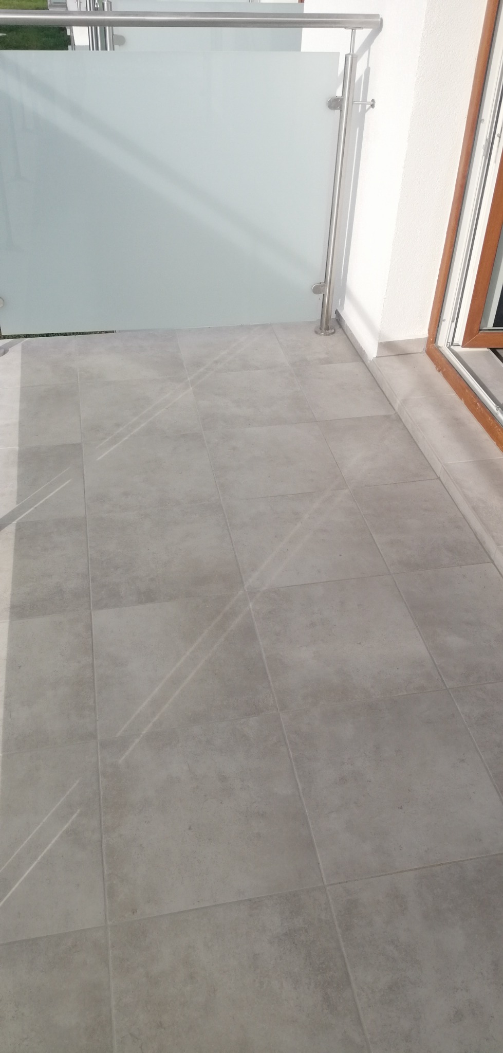 Wykończony balkon z szarymi płytkami podłogowymi, szklaną balustradą i metalowymi poręczami, widok z góry.