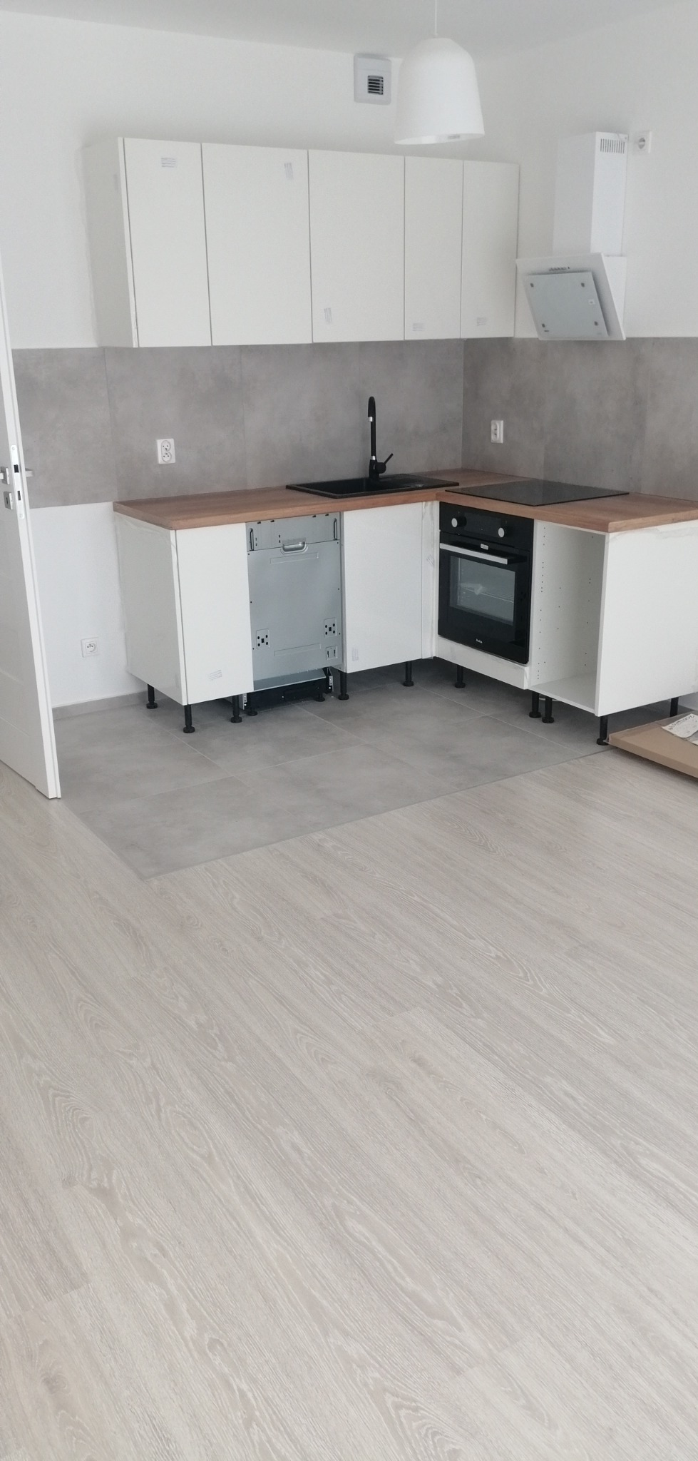 Wnętrze kuchni w trakcie montażu w Olsztynie: widoczne białe szafki kuchenne z drewnianymi blatami, czarny zlew i piekarnik, szare płytki na ścianie, jasne panele podłogowe i częściowo zamontowana...
