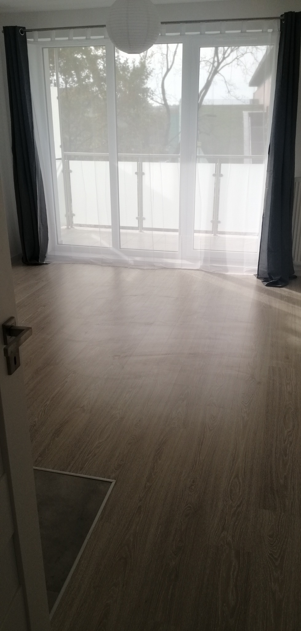 Jasne panele podłogowe w pokoju z balkonem, widoczne szare zasłony i białe firanki, lampa w kształcie kuli pod sufitem, częściowo widoczny próg i fragment drzwi.