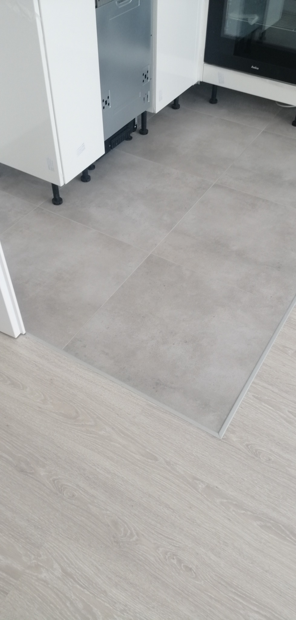 Połączenie jasnoszarych płytek podłogowych w kuchni z panelami podłogowymi w odcieniu jasnego drewna, widoczna listwa progowa.