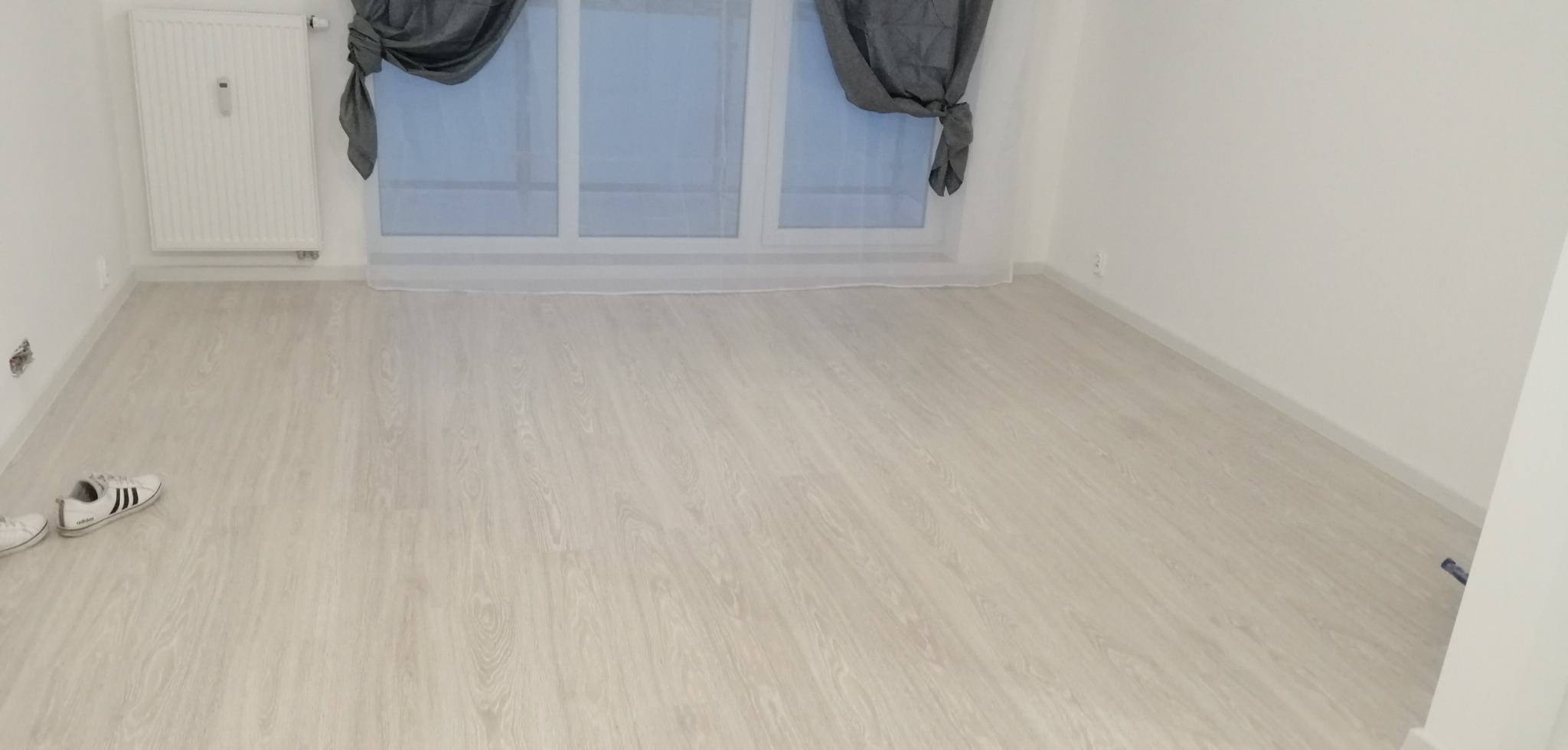 Puste, jasne pomieszczenie z panelami podłogowymi w odcieniach bieli i szarości, grzejnikiem na ścianie, oknem z szarymi zasłonami oraz parą butów sportowych w lewym dolnym rogu.