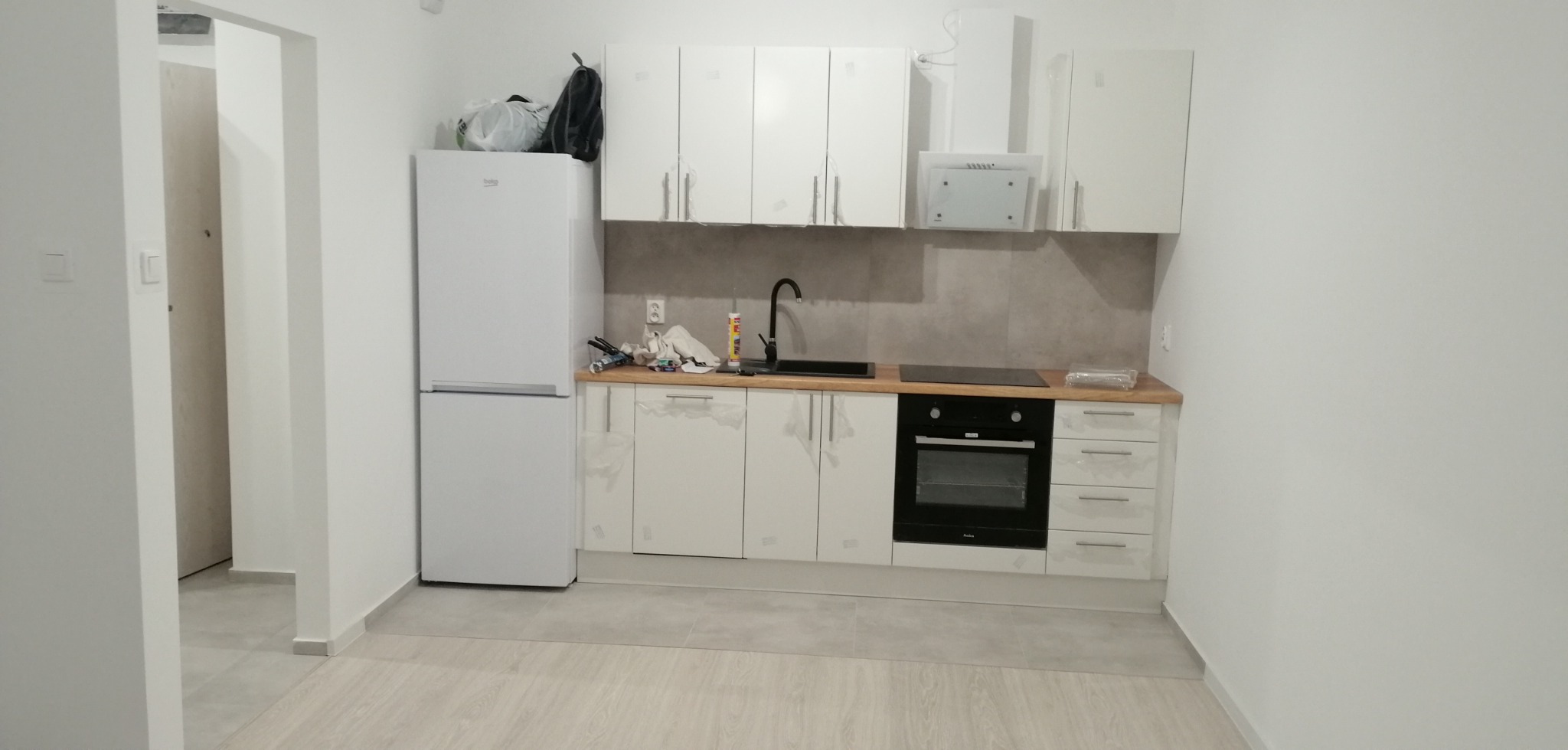 Nowoczesna kuchnia z białymi szafkami, czarnym zlewem i płytą indukcyjną, jeszcze z folią ochronną na frontach, widok na wejście do innego pomieszczenia.