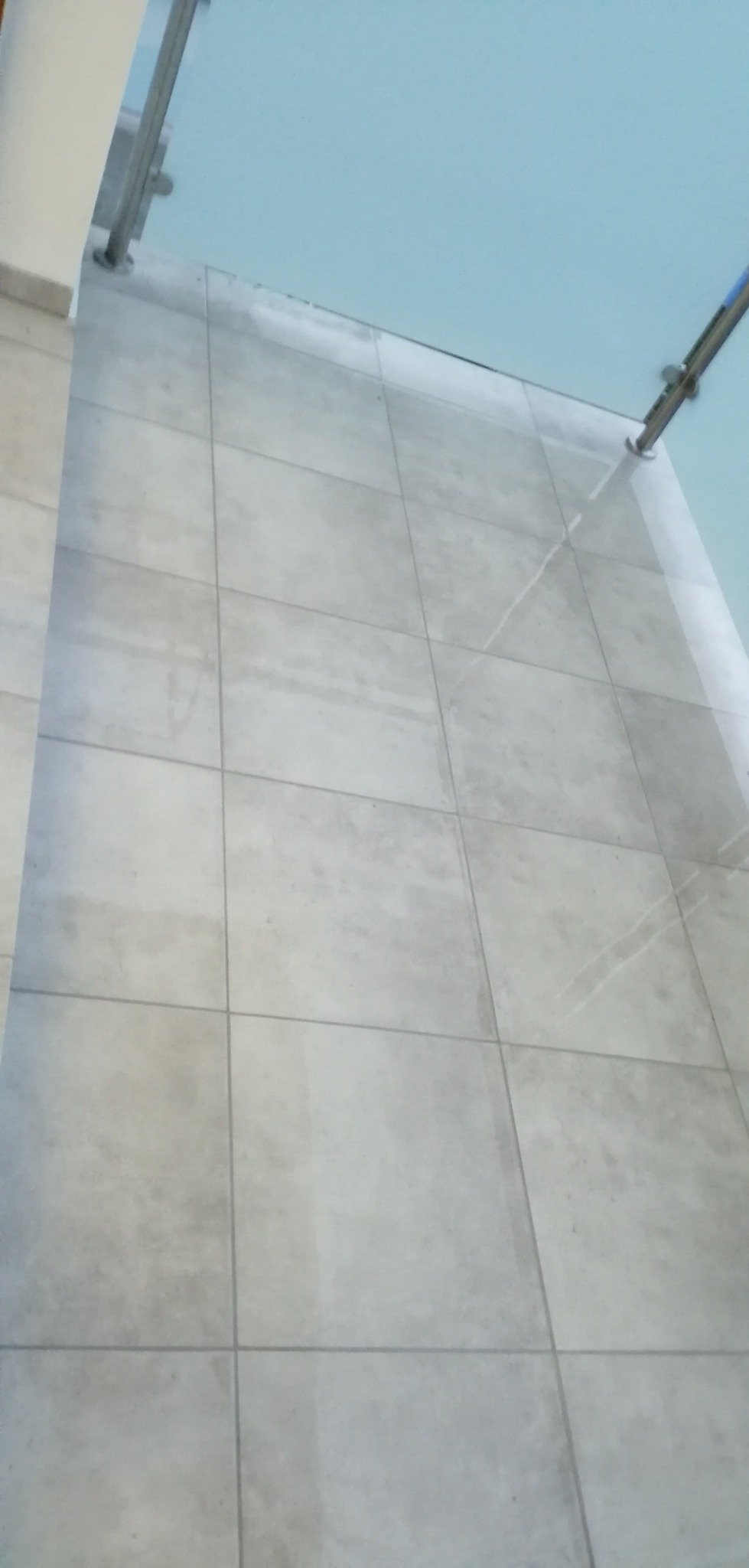 Jasnoszare płytki podłogowe ułożone w kwadraty z ciemniejszymi fugami, widoczne barierki balkonowe i niebieskie niebo odbijające się w szybie