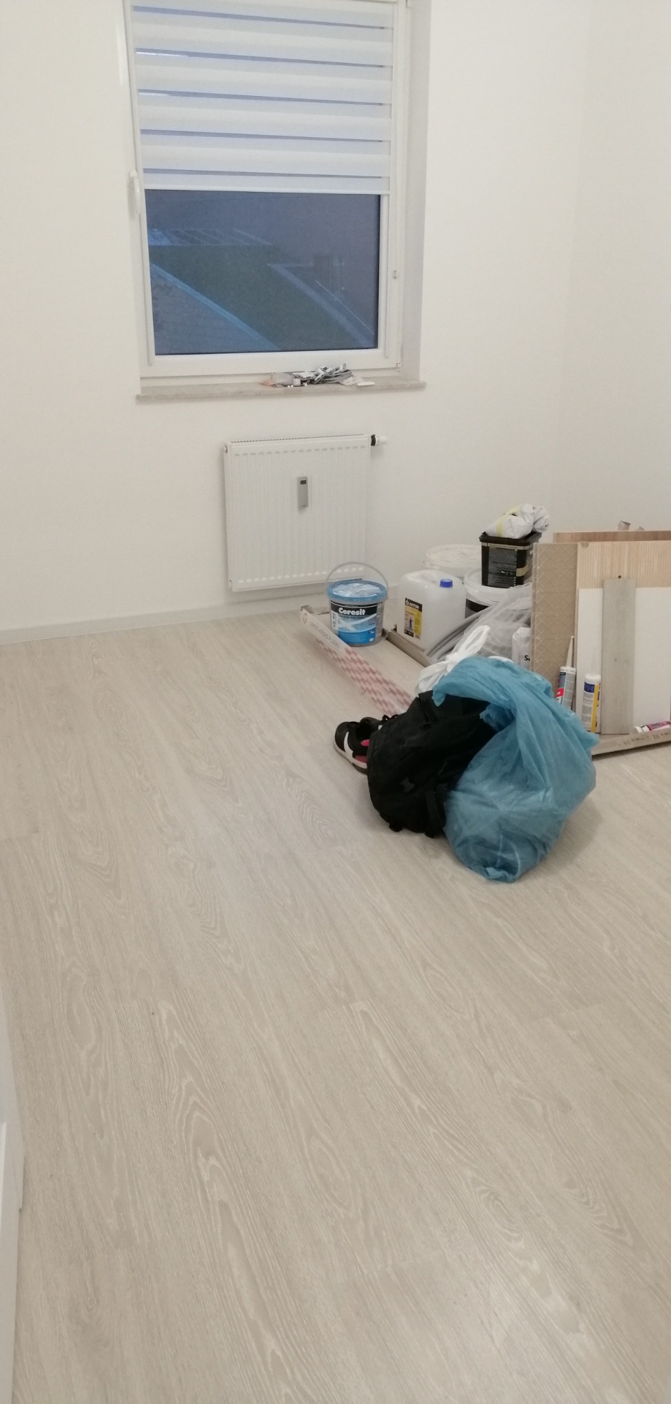 Pokój w trakcie prac wykończeniowych: jasne panele podłogowe, białe ściany, grzejnik, okno z roletą, wiadro z farbą, pojemniki, narzędzia oraz worek ze śmieciami.