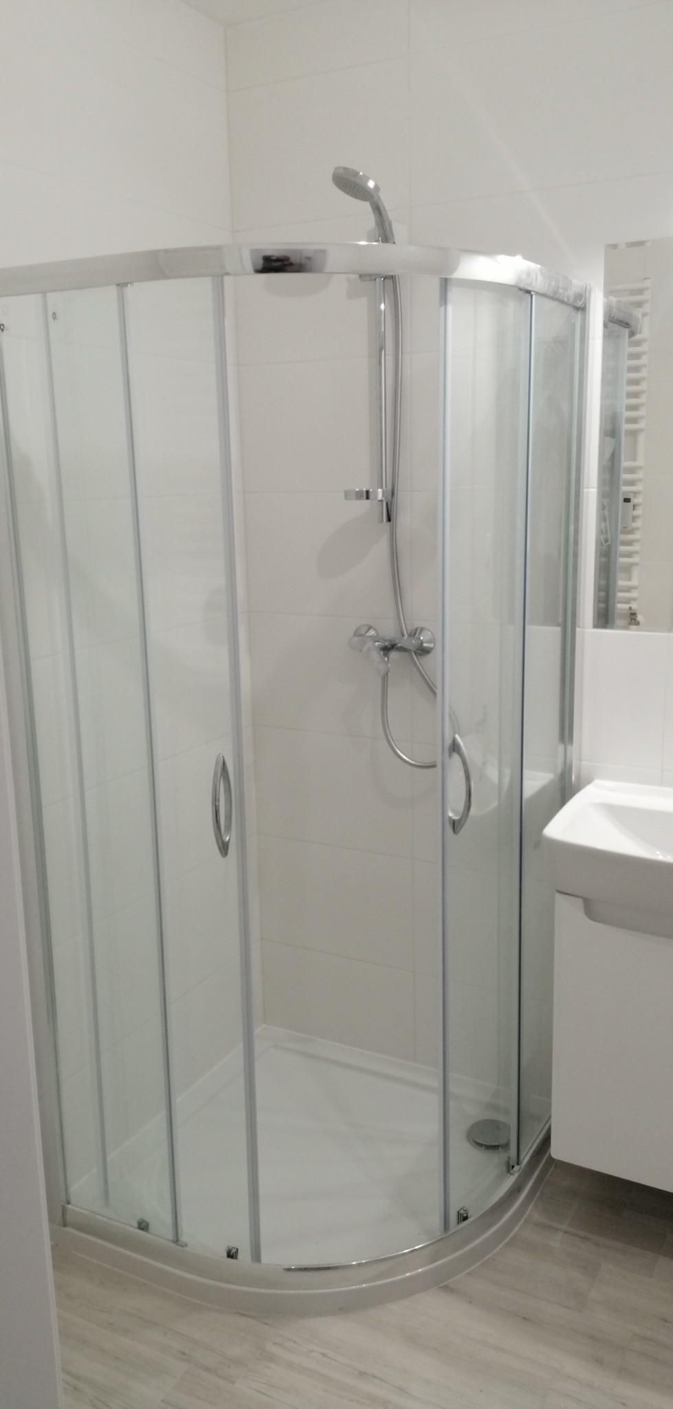 Nowoczesna kabina prysznicowa z chromowanymi profilami i przezroczystymi szybami, umywalka z szafką w tle, jasne płytki na ścianach i podłodze imitujące drewno.