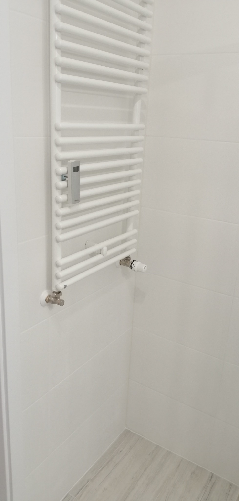 Biały, drabinkowy grzejnik łazienkowy z termostatem, zamontowany na ścianie z białych płytek, z widocznymi zaworami przyłączeniowymi i szarymi płytkami podłogowymi w rogu łazienki.