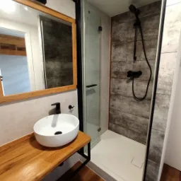 Nowoczesna łazienka z drewnianym blatem pod umywalką nablatową, czarną armaturą i kabiną prysznicową z grafitowymi płytkami imitującymi drewno na ścianie.