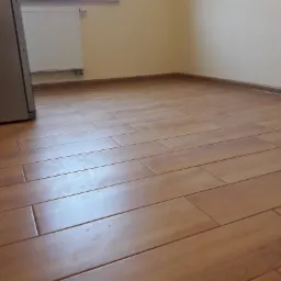 Ułożona podłoga z płytek imitujących drewno w pustym pokoju, widoczny fragment lodówki po lewej stronie.