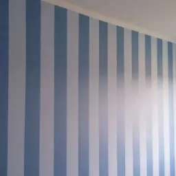 Ściana w pokoju pomalowana w pionowe, naprzemienne pasy w odcieniach błękitu i bieli, z widocznym oknem po prawej stronie i listwą przysufitową