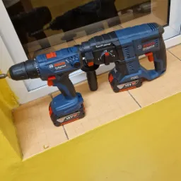 Dwa akumulatorowe wiertarko-wkrętarki Bosch Professional, w tym wiertarko-wkrętarka GSB 180-LI i młotowiertarka GBH 18V-26, leżące na brązowych płytkach przy żółtej ścianie i oknie.