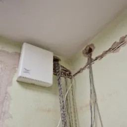 Biała centrala alarmowa zamontowana na zielonej ścianie, z widocznymi kablami wychodzącymi z otworu w ścianie i biegnącymi wzdłuż listwy przypodłogowej