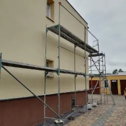 Budynek z beżową elewacją w trakcie prac, widoczne rusztowanie, okna zabezpieczone folią, dolna część elewacji w brązowym kolorze, kostka brukowa na podwórzu.